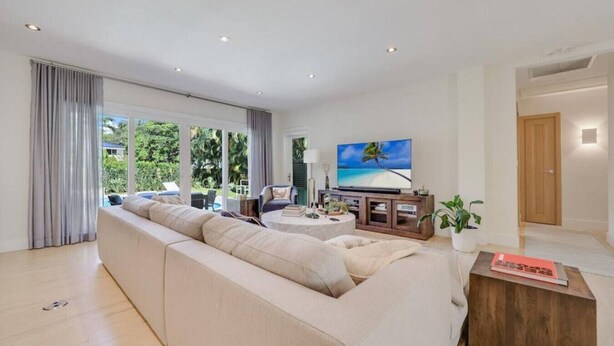 A modern living room with a large beige sofa, a coffee table, and a flat-screen TV. There is a sliding glass door leading to a balcony with a pool. - High-End House with Heated Pool, outdoor TV (Miami Shores)
