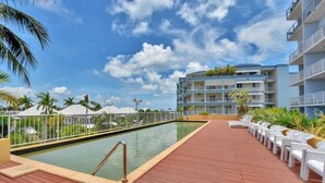 Uma área de piscina com deck de madeira, cadeiras de descanso e um edifício com varandas ao fundo.