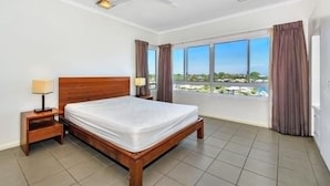 Quarto de hotel com cama de madeira, mesa de cabeceira, ventoinha no teto e uma grande janela com cortinas.