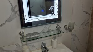 Une salle de bain avec un lavabo blanc, une étagère en verre, un miroir avec un cadre décoratif et des murs en marbre.