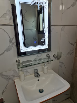 A bathroom with a white sink, a glass shelf, a mirror with a decorative frame, and marble walls. - STUDIO HAPPY HOME (Dakar)