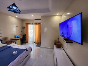 A hotel room with a bed, a desk with a laptop, a TV on the wall, and a ceiling light fixture. - STUDIO HAPPY HOME (Dakar)