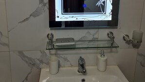Une salle de bain avec un pare-baignoire en marbre, une étagère en verre au-dessus du lavabo et un miroir doté d’un cadre décoratif.