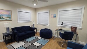 Un salon avec un canapé bleu foncé, une table basse en verre, un fauteuil en forme de poire bleu et une chaise de bureau en verre. Il y a trois fenêtres avec des stores blancs, un ventilateur de plafond et un tableau au mur.