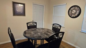 Un coin repas avec une table ronde et quatre chaises, un tableau encadré au mur, une porte et une fenêtre avec des stores.