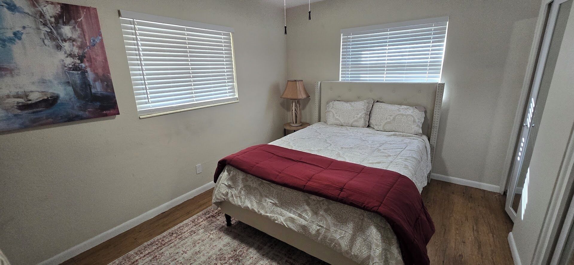 A bedroom with a bed, two windows with blinds, a nightstand with a lamp, and a painting on the wall.