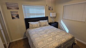 Une chambre à coucher avec un lit, une table de chevet, une lampe et deux fenêtres équipées de stores.