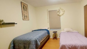 Une petite pièce simple, avec deux lits simples, une petite table, une fenêtre avec un store et une armoire en bois.