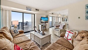 A spacious living room with a large TV, comfortable seating, and a balcony with a view. - 2 BR Condo w/ Sunrise and Sunset Views (Ocean City)