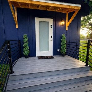 Une terrasse en bois avec une porte blanche, des rampes noires et deux plantes en pot.