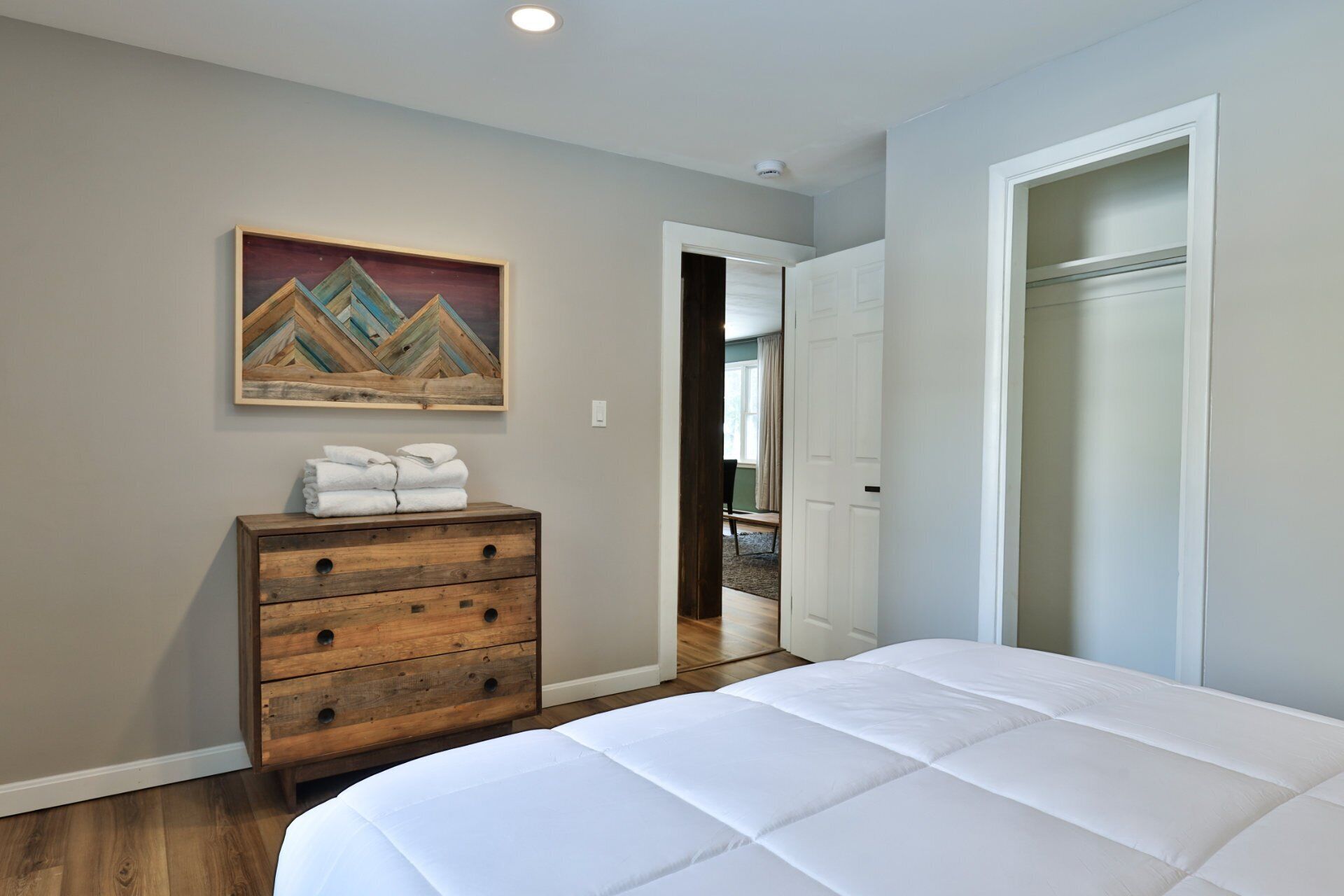 A bedroom with a bed, a wooden dresser, a painting, and a door leading to another room.
