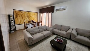 Una sala de estar con un sofá modular, una mesa de centro, una mesa de comedor con sillas, un aire acondicionado instalado en la pared y un cuadro abstracto de gran tamaño.