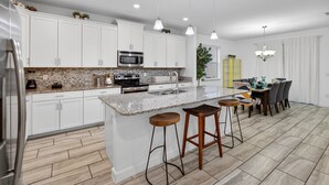 Une cuisine moderne avec des armoires blanches, un comptoir en granit, un réfrigérateur en acier inoxydable, un four à micro-ondes et un évier. On y trouve trois tabourets de bar en bois et un coin repas avec une table et des chaises.