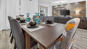 Un espace de repas moderne avec une table en bois dressée pour deux personnes, entourée de chaises à revêtement gris. Un lustre est suspendu au plafond et un téléviseur à écran plat est fixé au mur.