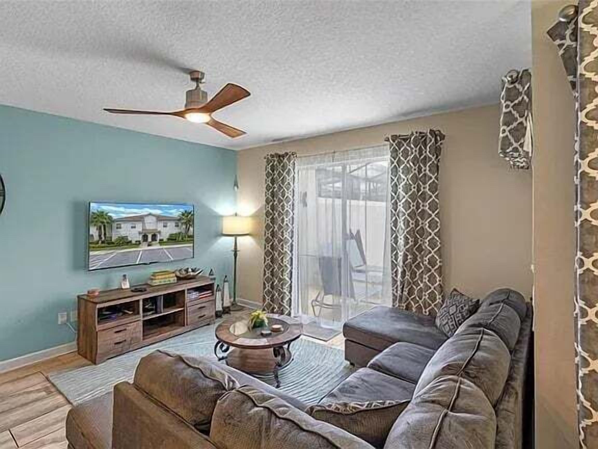 A living room with a sectional sofa, a coffee table, a TV on a stand, a ceiling fan, and a window with patterned curtains.