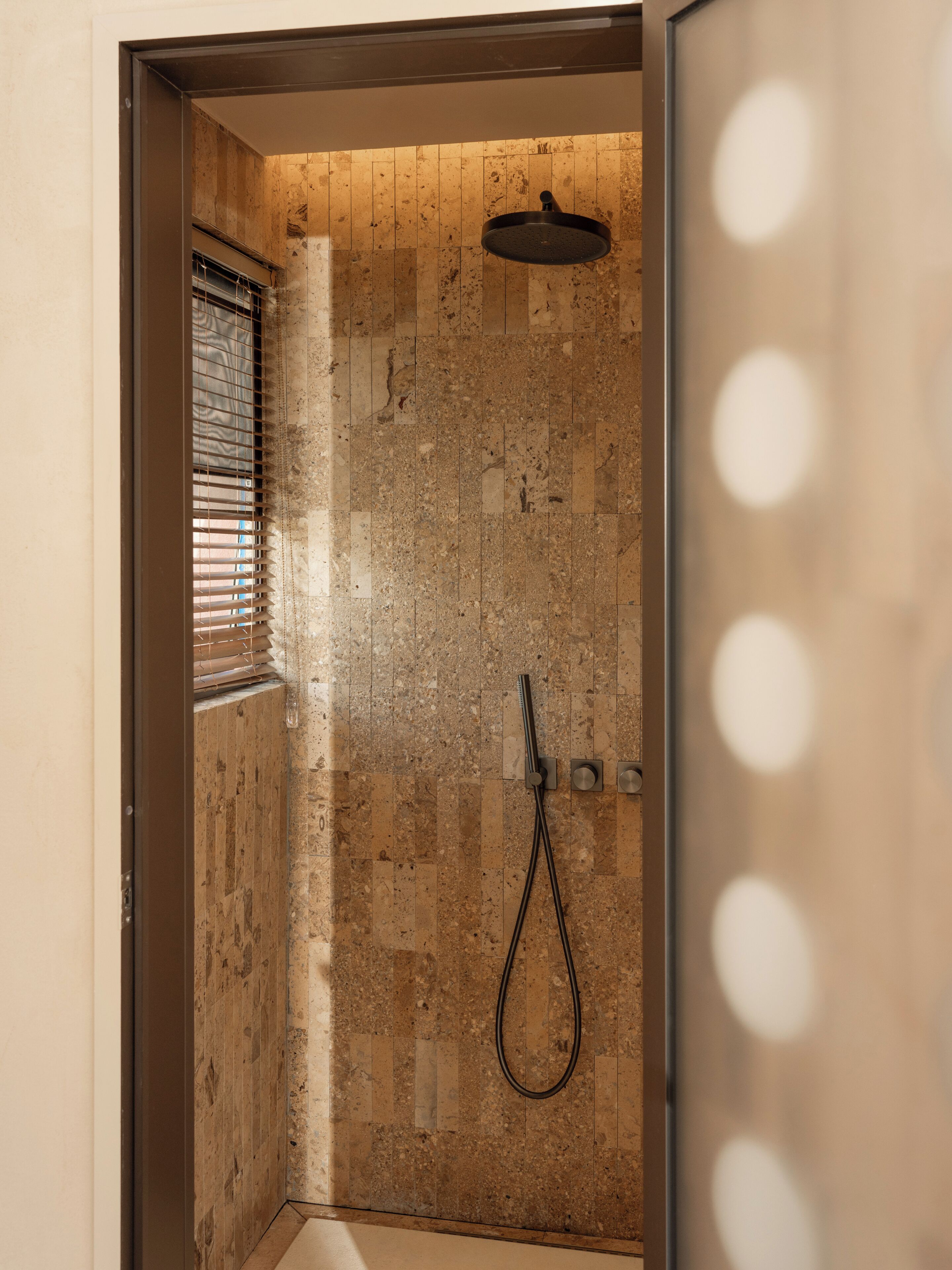 A shower area with a tiled wall, a showerhead, and a window with blinds.