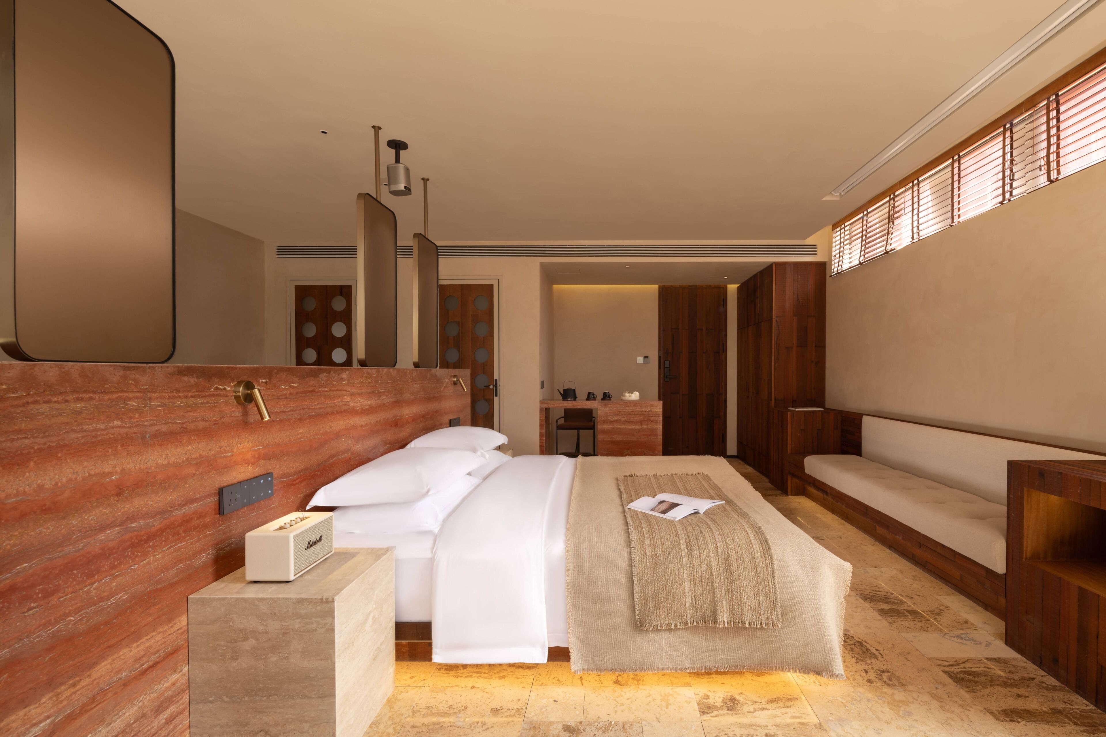 A modern hotel room with a large bed, a wooden headboard, a bedside table with a lamp, and a view of the hallway.
