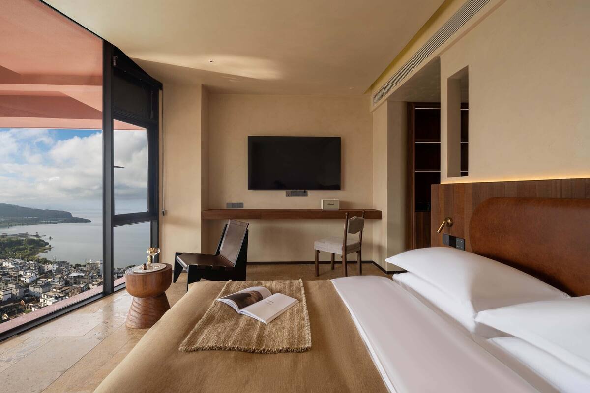 A hotel room with a large window, a bed, a desk with a chair, a television, and a view of a city and water.