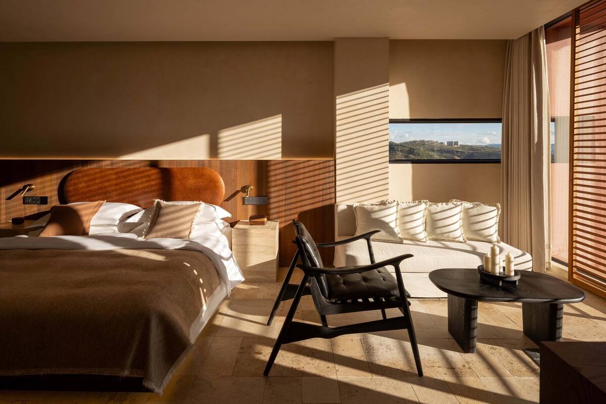 A modern hotel room with a large bed, a sofa, a chair, a small table, and a view of the outdoors.