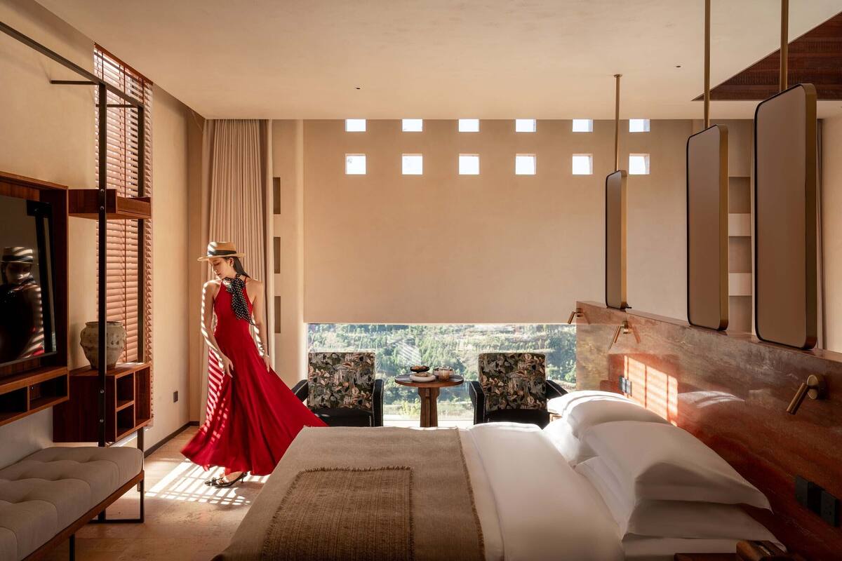 A modern hotel room with a large bed, a small table with a vase, and a view of the outdoors.