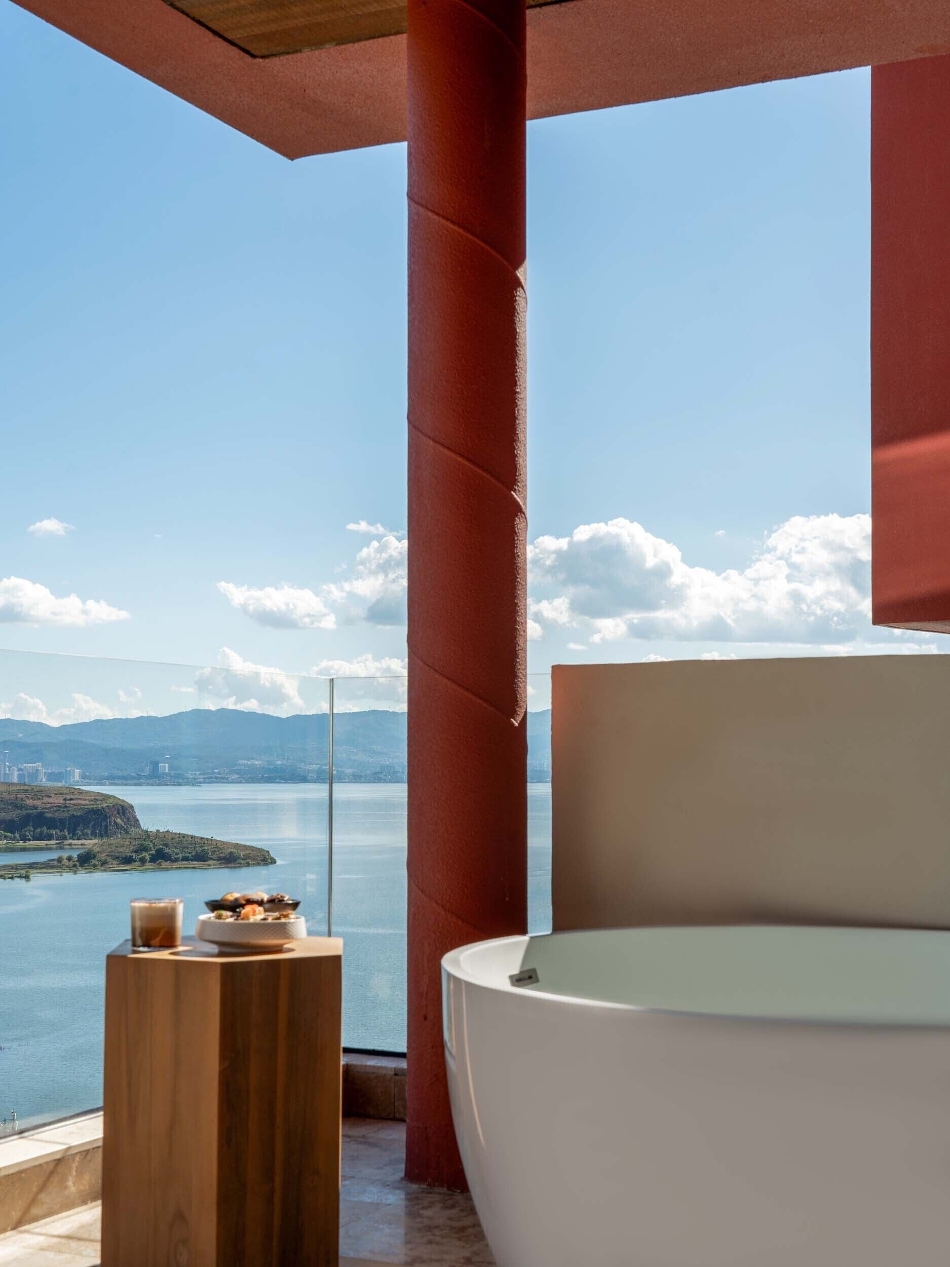Une salle de bain moderne avec une grande baignoire, une table d’appoint en bois sur laquelle se trouve un verre d’eau, et une vue sur un lac avec des montagnes en arrière-plan.