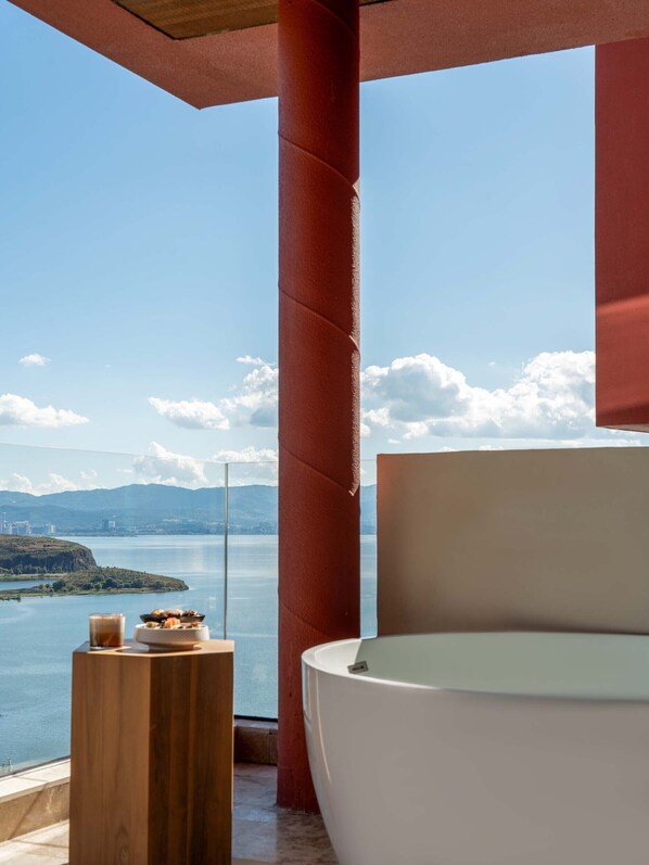 Une salle de bain moderne avec une grande baignoire, une table d’appoint en bois sur laquelle se trouve un verre d’eau, et une vue sur un lac avec des montagnes en arrière-plan.