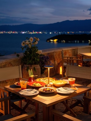 Un espace de restauration en plein air, avec une table dressée pour un repas, offrant une vue sur une étendue d’eau et les lumières lointaines de la ville.