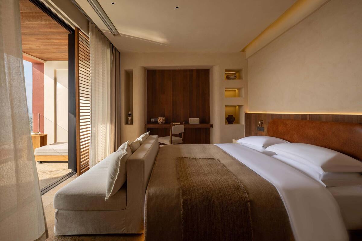 A modern hotel room with a large bed, a sofa, a desk, and a view through a sliding door.