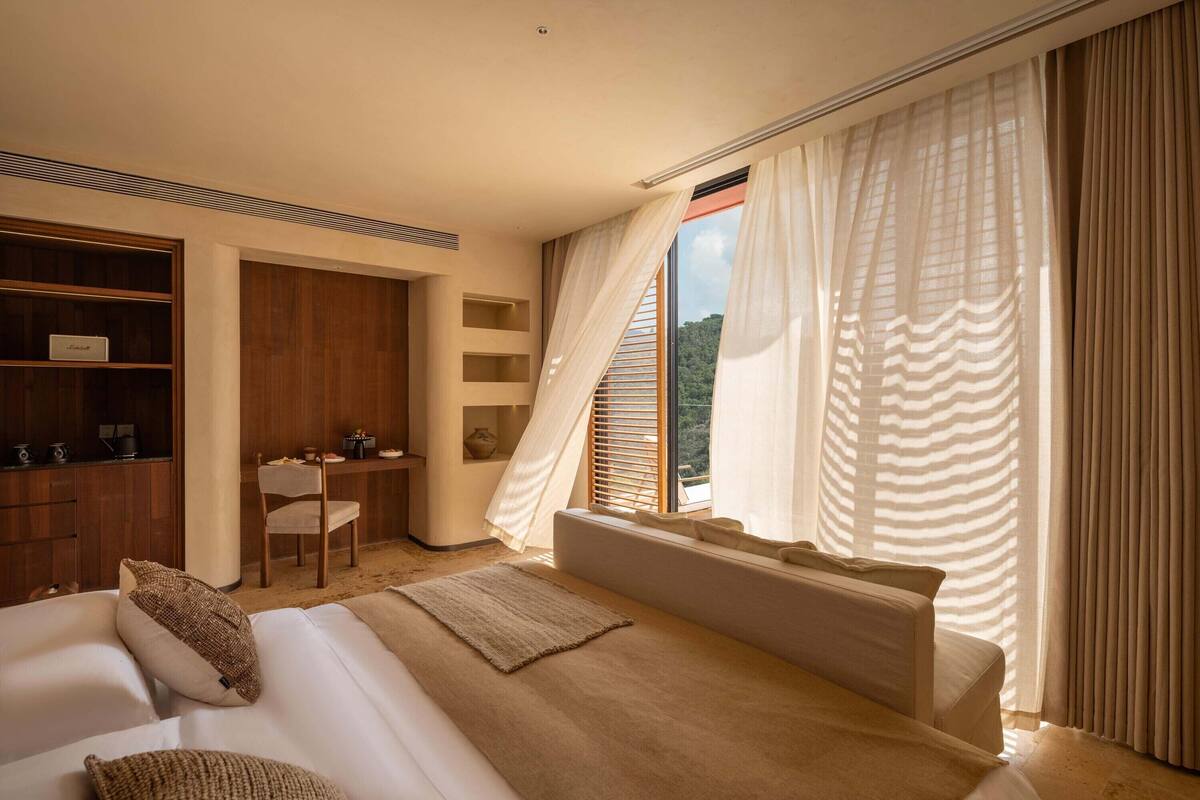 A bedroom with a large bed, a desk, and a chair. There is a view of the outdoors through the window.