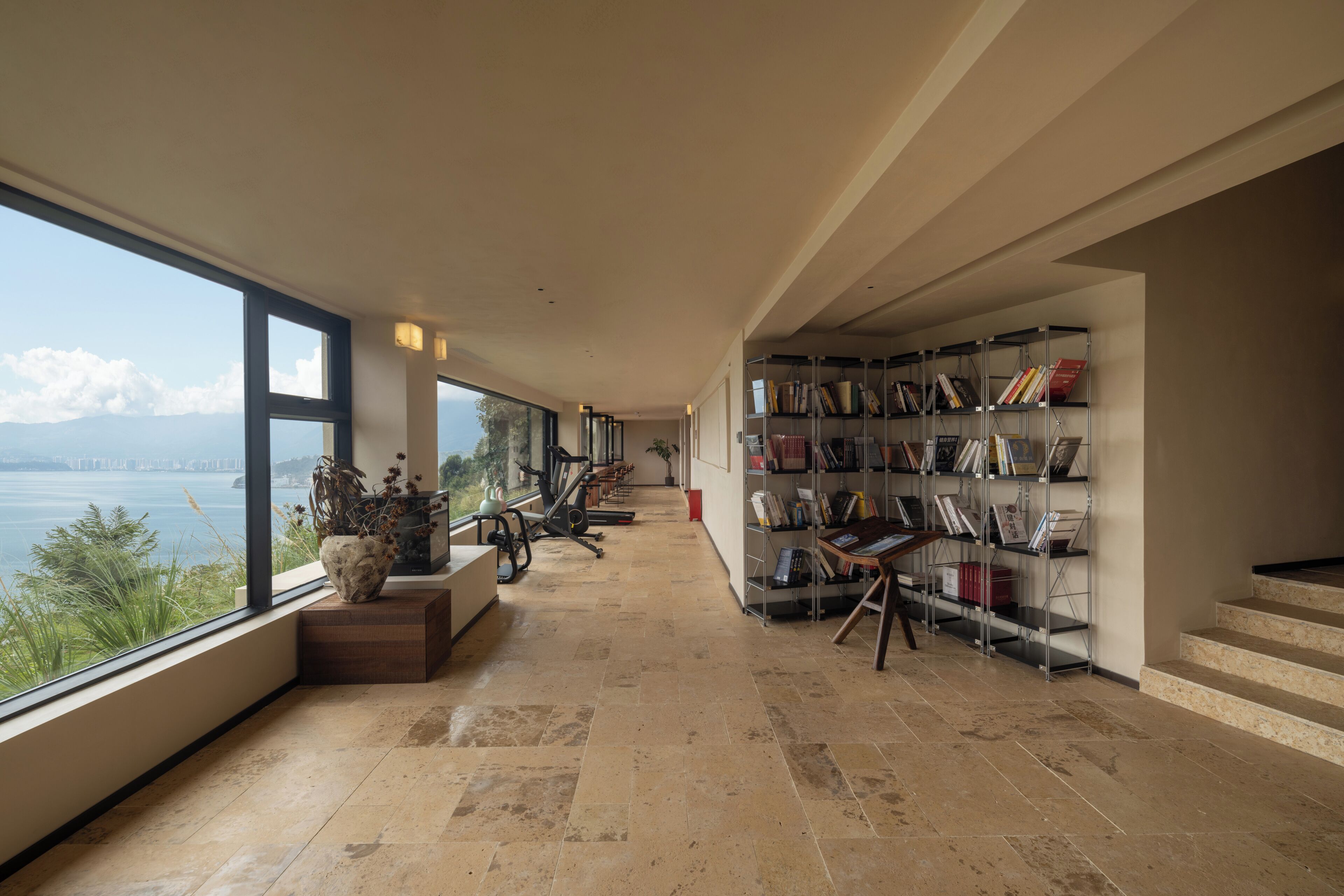 A spacious hallway with a large window overlooking a body of water, a treadmill, and a bookshelf filled with books.