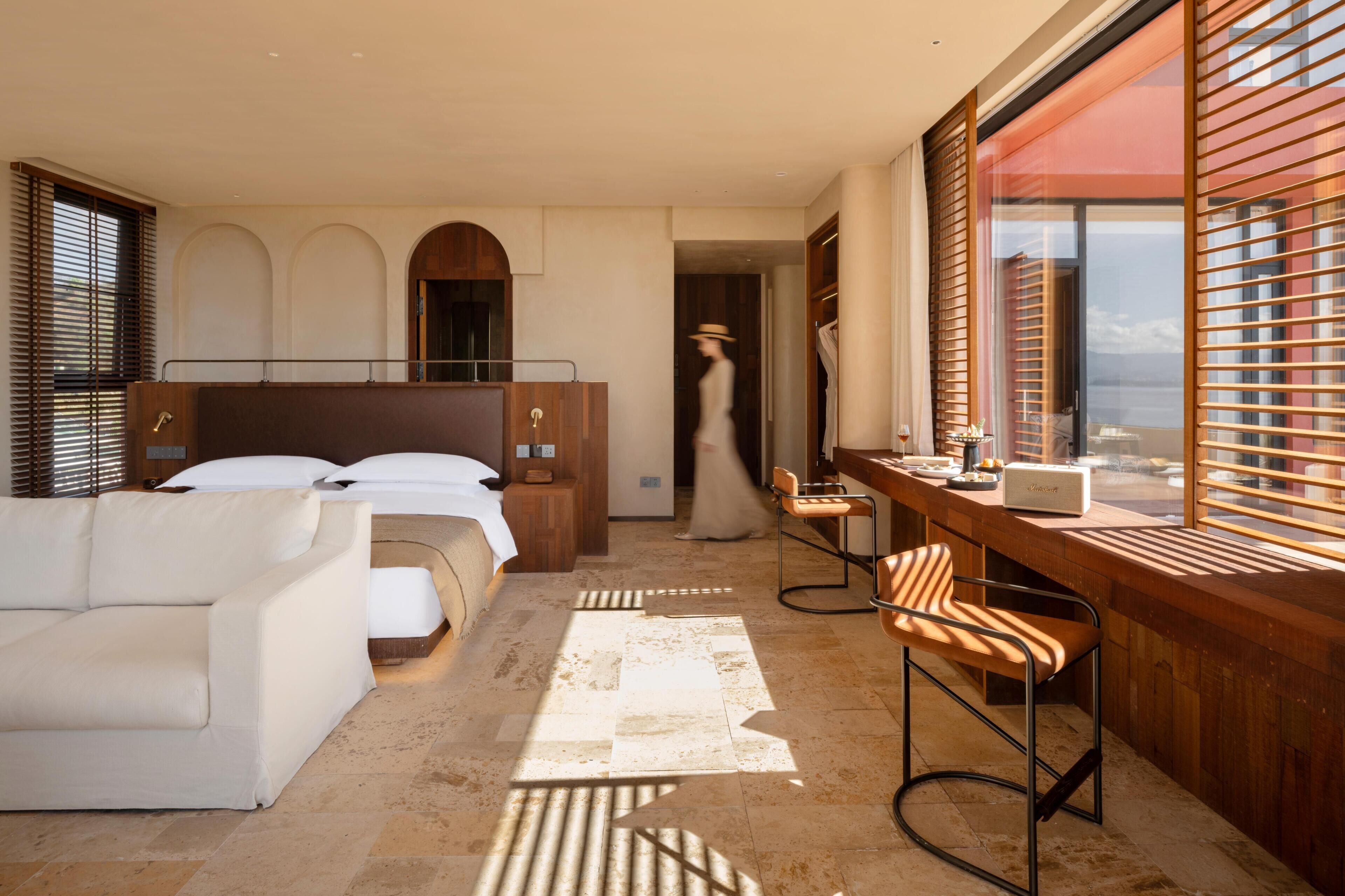 A hotel room with a bed, a sofa, a desk with a chair, and a view through the window.