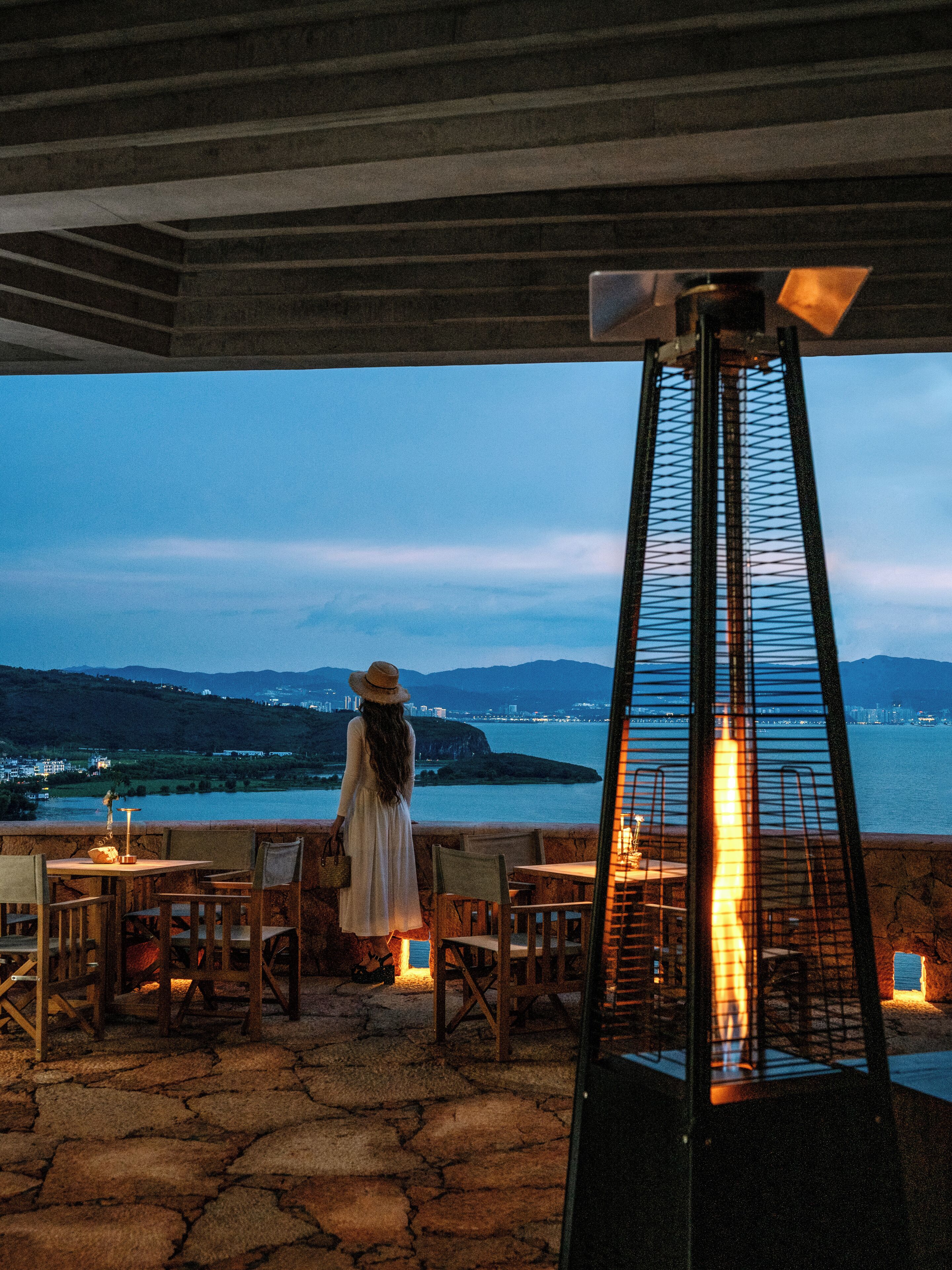 Una terraza con vistas a un cuerpo de agua y colinas distantes, con mobiliario exterior y una chimenea central.