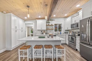 Una cocina moderna con armarios blancos, una isla central con taburetes, electrodomésticos de acero inoxidable y vigas de madera en el techo.