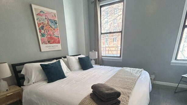 A neatly made bed with a white comforter and two pillows, a bedside table with a lamp, and a window with a view of buildings.