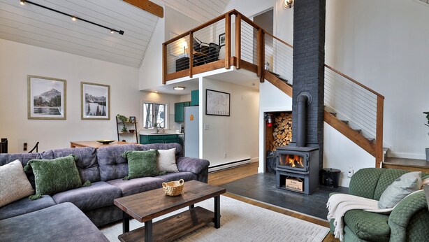 Ein geräumiges Wohnzimmer mit einem großen Ecksofa, einem Holztisch und einem Kamin. Eine Treppe führt zu einer oberen Ebene mit einem Balkon.