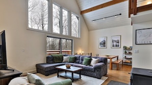 Ein geräumiges Wohnzimmer mit einer Eckcouch, einem Couchtisch und einem Kamin. Große Fenster sorgen für Tageslicht und Blick ins Grüne.