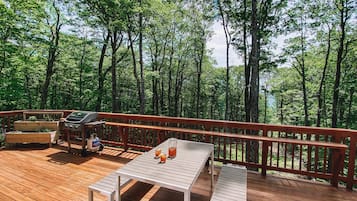 A wooden deck with a grill, table, and benches, surrounded by a forest.