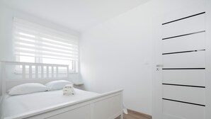 A white bedroom with a bed, a window with blinds, a bedside table, and a wardrobe with horizontal lines.
