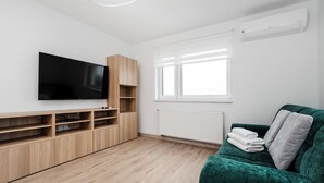 A modern living room with a flat-screen TV, wooden shelving, a green sofa, and a window with blinds.