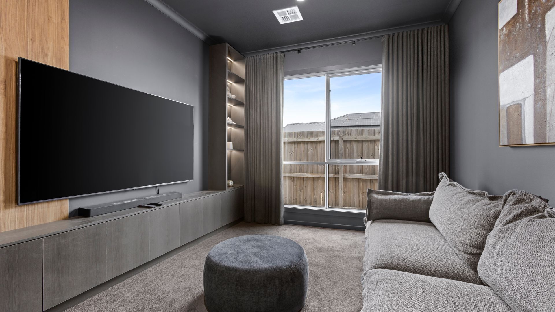 A modern living room with a large flat-screen TV mounted on a wooden panel, a grey sofa, a grey ottoman, and a large window with curtains.