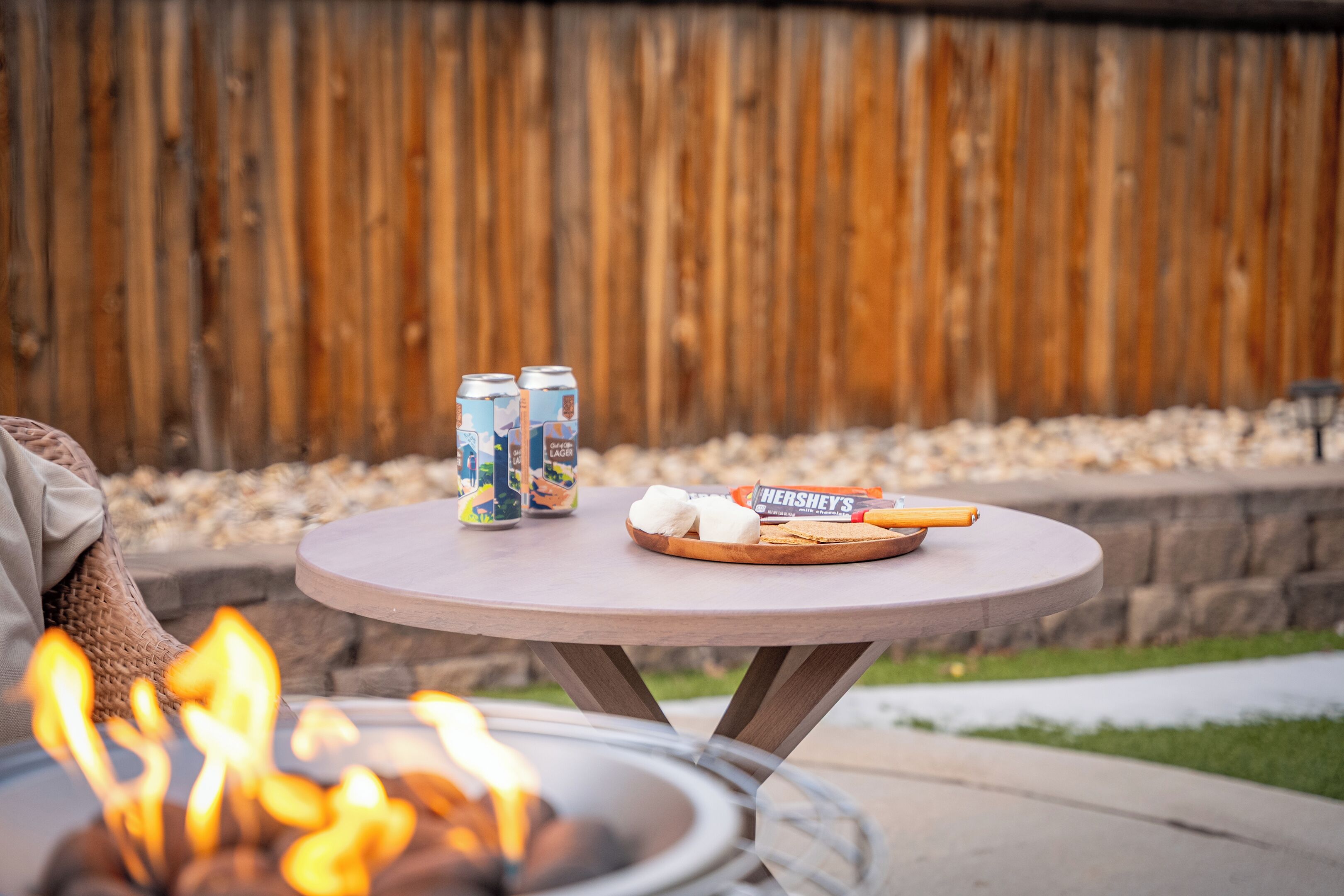 A round table with snacks and drinks, a fire pit in the foreground, and a wooden fence in the background.