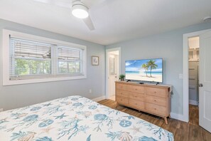 Un dormitorio con una cama, un televisor sobre una cómoda, un ventilador de techo y una puerta que da a un baño.