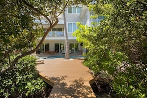 A wooden deck leading to a pool area with lounge chairs and a building in the background. - A Shore Thing: On Canal! Pool Spa!  Rooftop Deck by Anna Maria Life Vacation Rentals (Anna Maria)