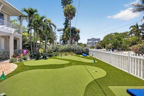 A well-maintained artificial grass golf course with a white picket fence, surrounded by lush tropical vegetation and residential buildings. - A Shore Thing: On Canal! Pool Spa!  Rooftop Deck by Anna Maria Life Vacation Rentals (Anna Maria)