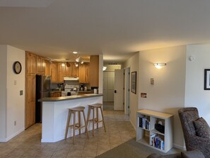 A kitchen with wooden cabinets, a central island with stools, a refrigerator, and a dining area with a bookshelf. - Downtown Bayfield Condo 204 @ Washington Ave (Bayfield)