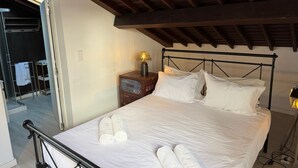 Una cama bien hecha con sábanas blancas y toallas enrolladas, un cabecero de metal, una mesita de noche con una lámpara y vistas a un baño con un perchero para toallas.