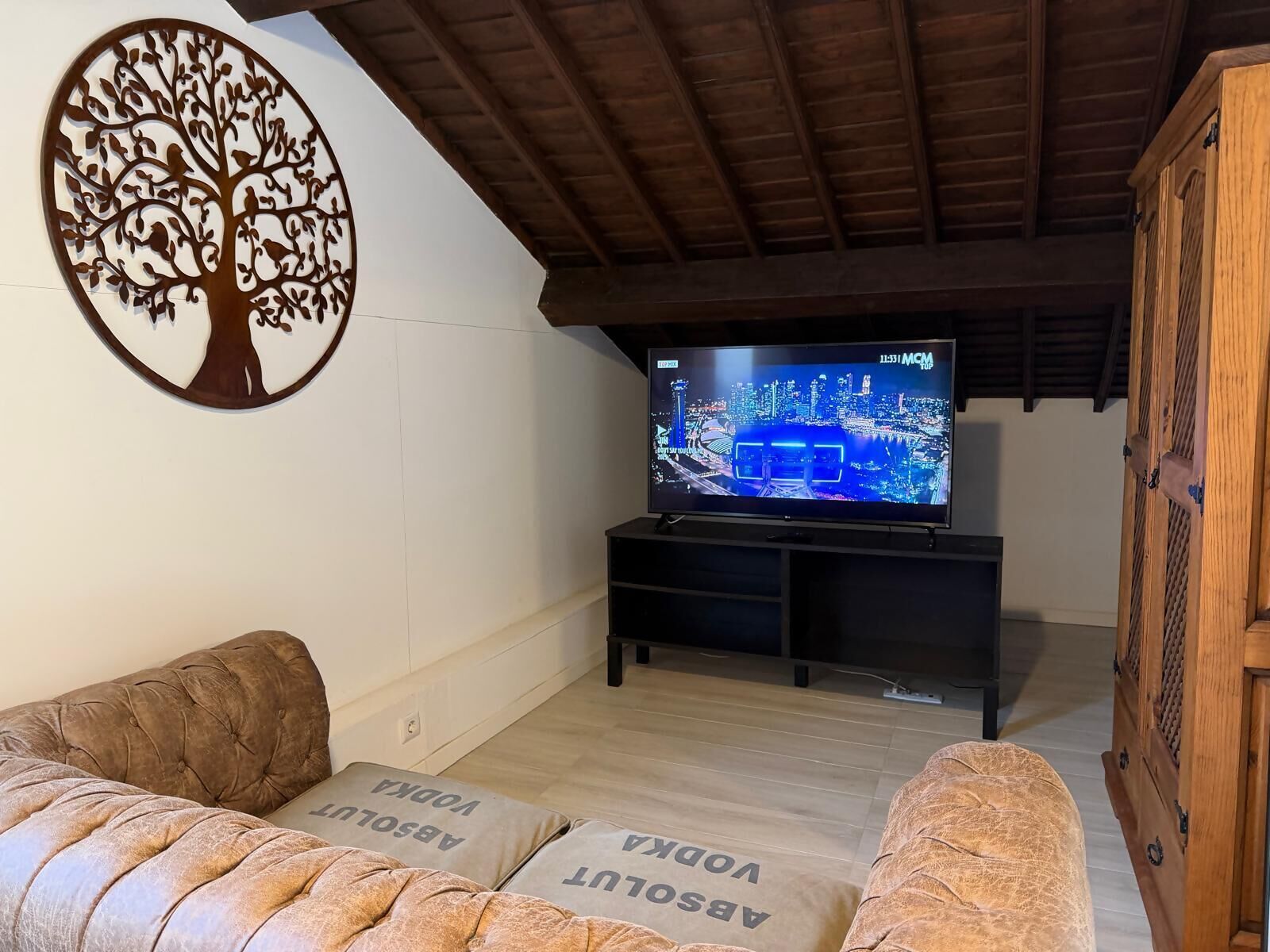Una sala de estar con una televisión mostrando un paisaje urbano, techo de madera y un adorno mural con forma de árbol.