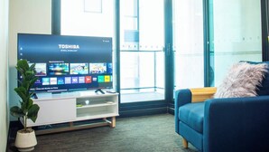 Olohuoneessa on litteänäyttöinen televisio, josta näkyy erilaisia suoratoistopalveluita, ruukussa oleva kasvi, sininen sohva ja lasiliukuovi, joka johtaa parvekkeelle.
