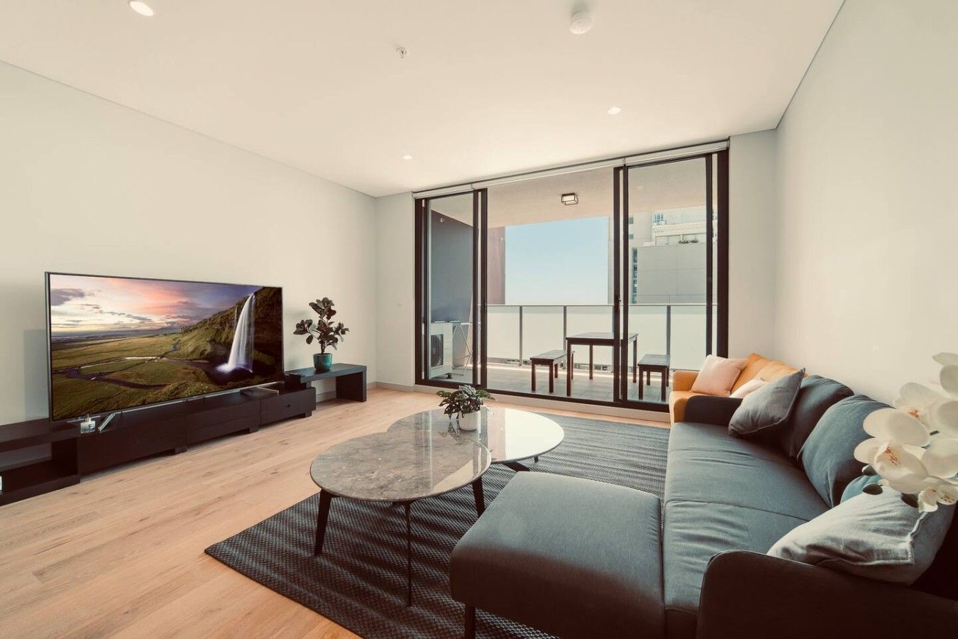 A modern living room with a flat-screen TV, a sofa, a coffee table, and a balcony with outdoor seating.