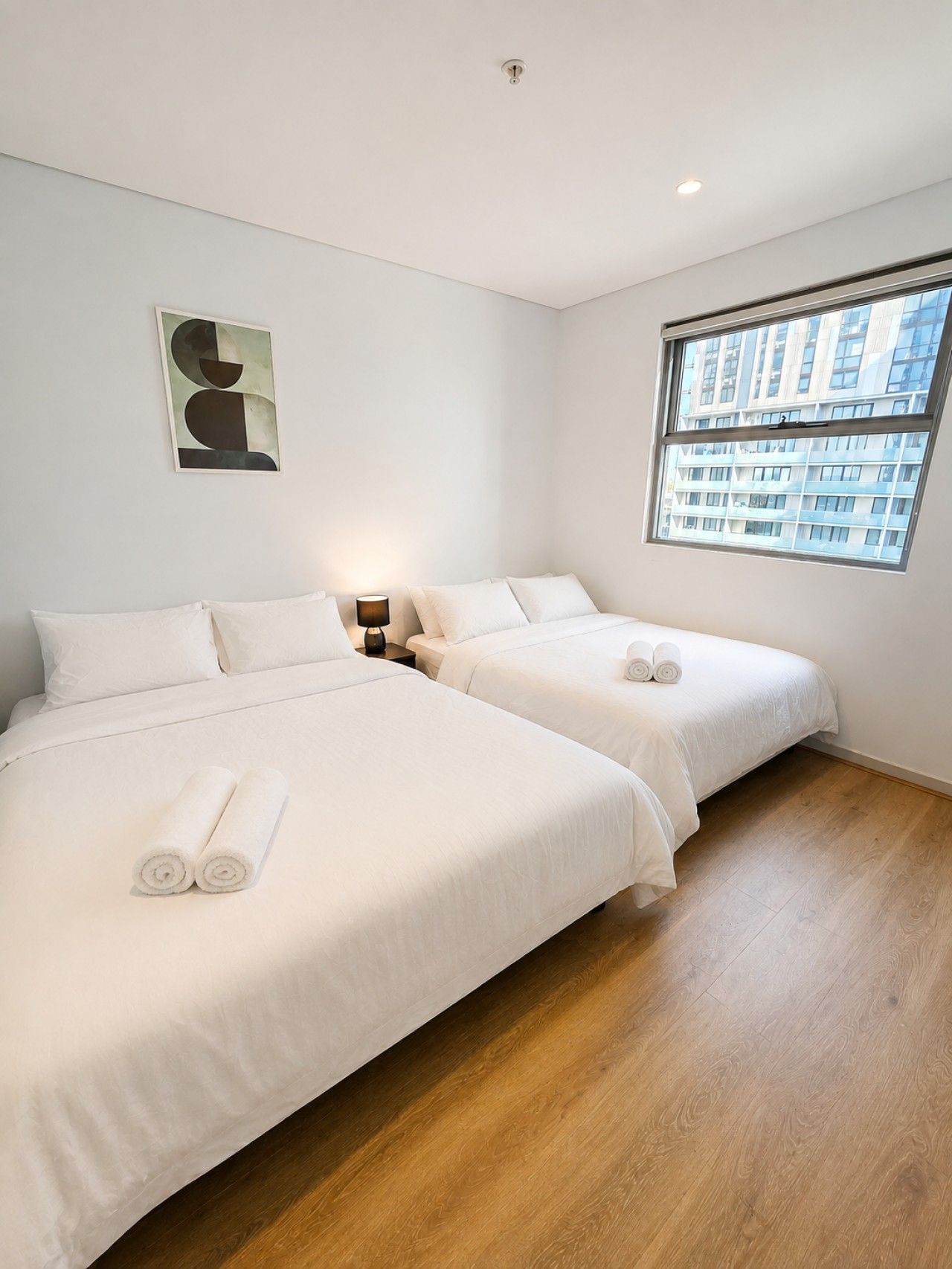 Een hotelkamer met twee bedden, een nachtkastje, een lamp, een schilderij en een raam met uitzicht op gebouwen.